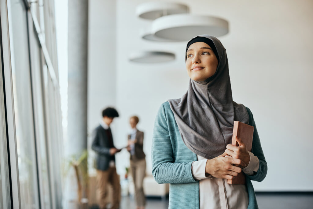 Smiling Muslim businesswoman standing in hallway at corporate office and looking through the window.